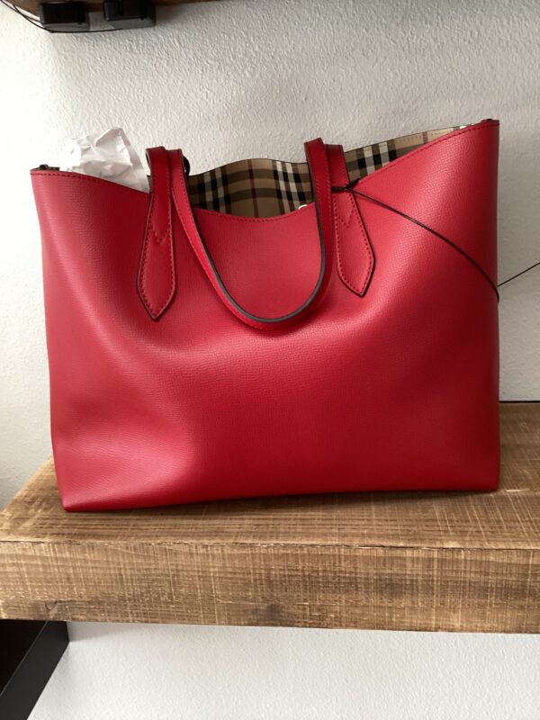 A Burberry red reversible tote bag with plaid interior lining, resting on a wooden shelf against a white wall.