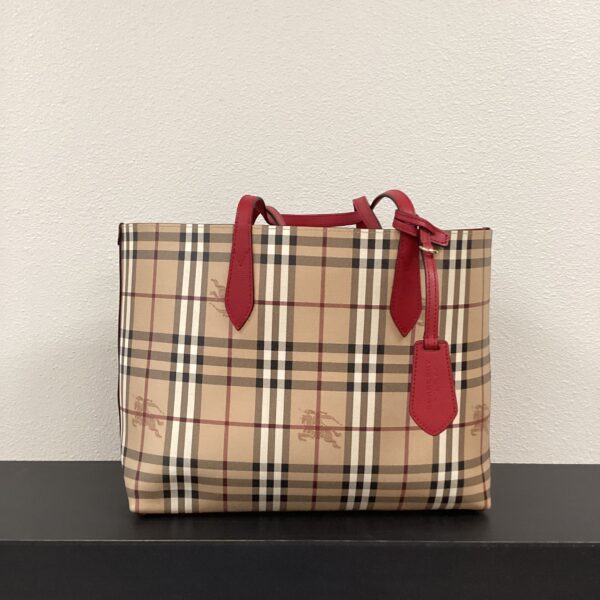 Beige plaid Burberry tote bag with red handles and accents, displayed on a white shelf against a gray wall.