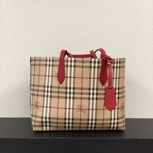 A Burberry red reversible tote bag with beige plaid pattern, displayed on a white surface against a grey wall.
