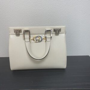A cream-colored designer handbag with a gold ring clasp and dual handles, displayed on a black shelf against a gray wall.