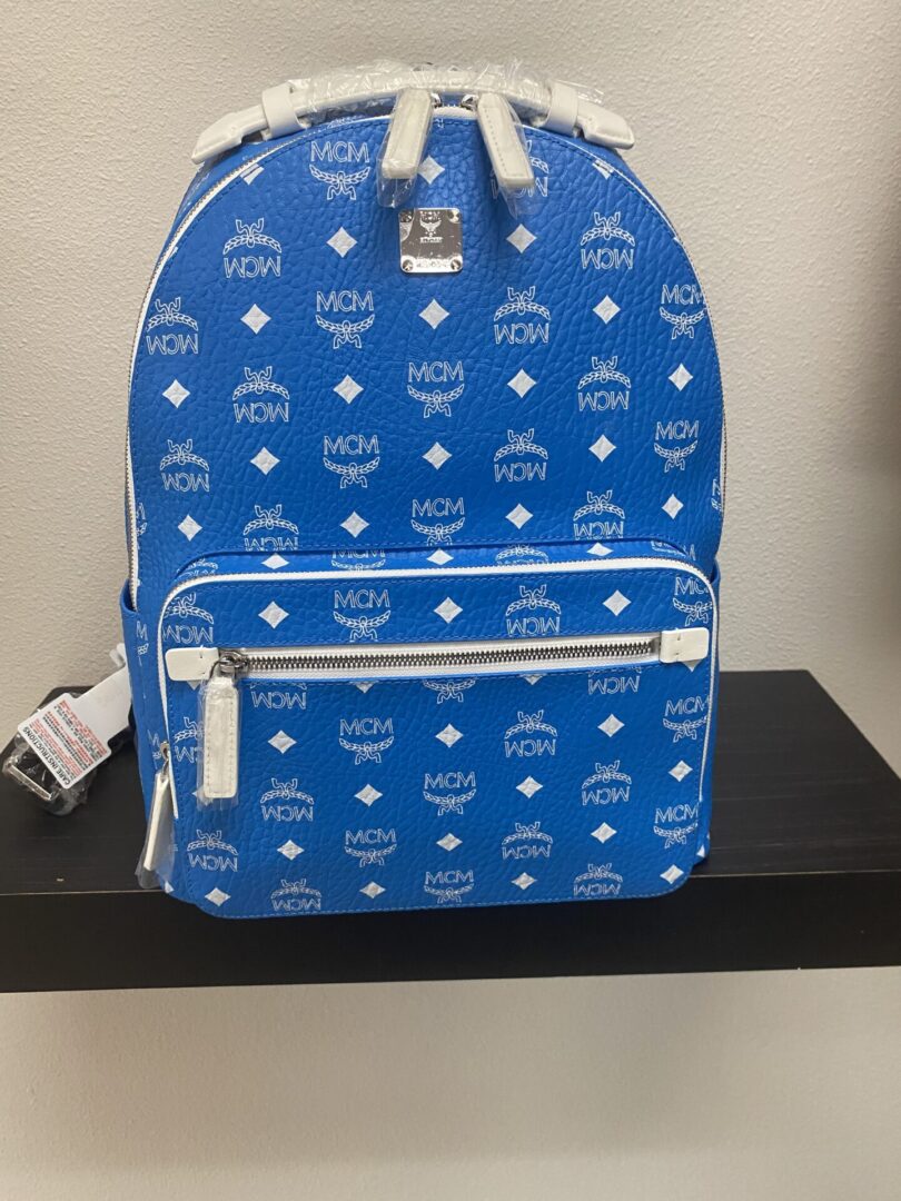 Blue mcm branded backpack with logo pattern, featuring a top handle, zippered compartments, and a price tag, displayed on a black shelf.