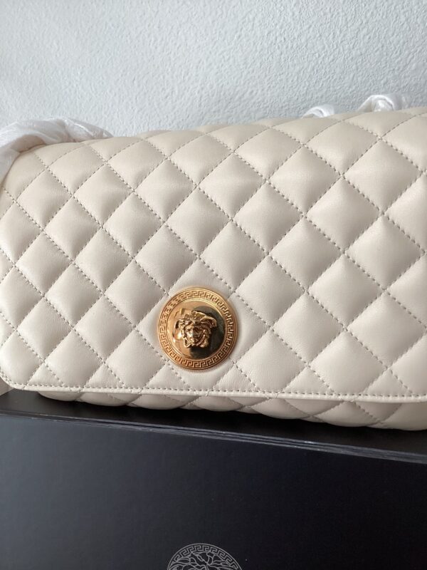 Close-up of a white quilted clutch with a golden circular logo featuring a lion's head, resting on a black box.