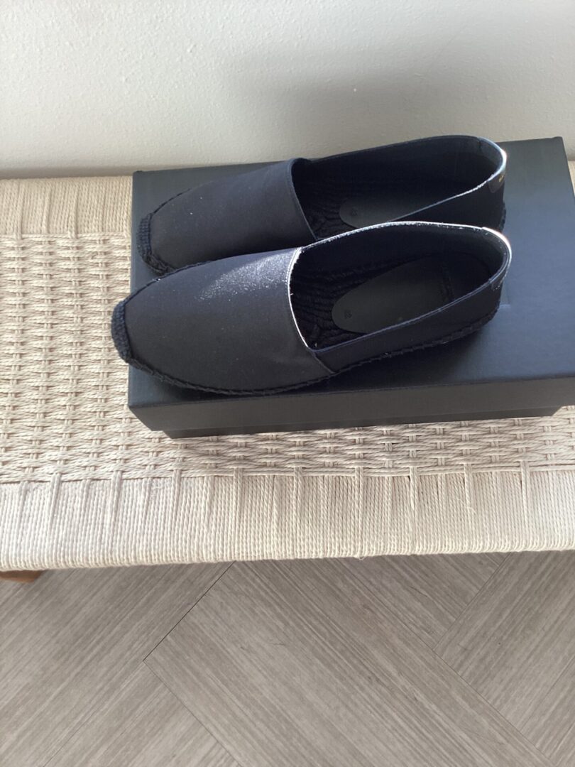 A pair of YSL Espadrilles placed neatly on a gray shoebox, on top of a woven beige mat on a wooden floor.