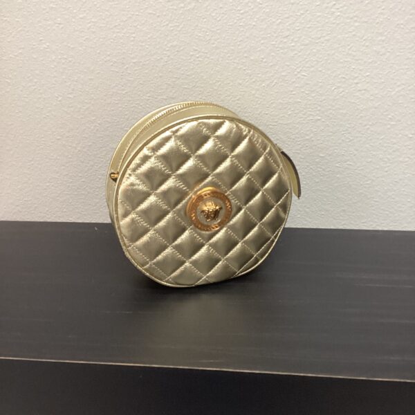 A small, round, metallic gold VERSACE purse with quilted stitching and a central logo, displayed on a gray surface against a blue wall.