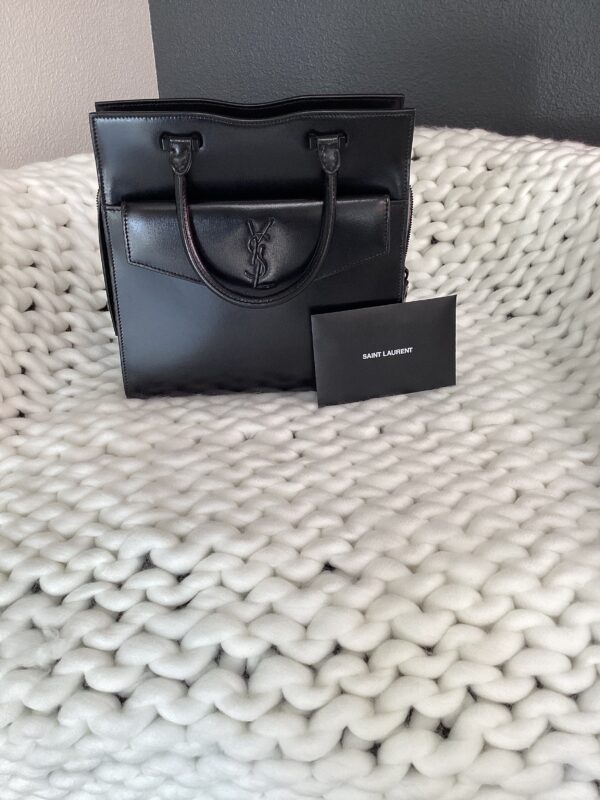 A black YSL Uptown Bag resting on a textured white blanket, accompanied by a brand card.