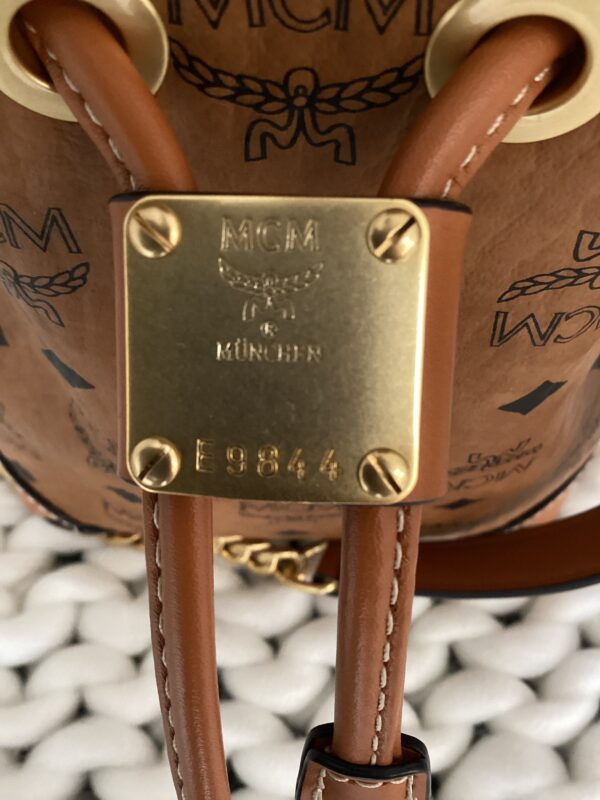 Close-up of a tan MCM Bucket Bag showing a detailed view of the brand's gold logo plate and part of the leather straps.
