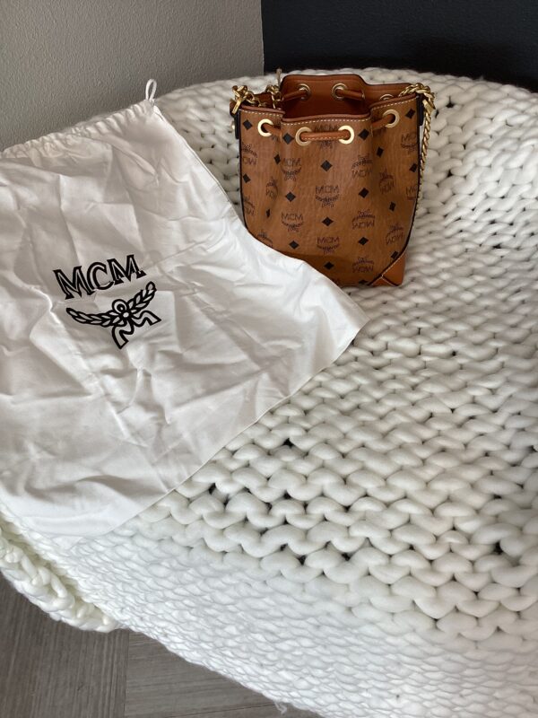 A small, brown MCM Bucket Bag with logo pattern and a matching white drawstring dust bag, placed on a textured white blanket.