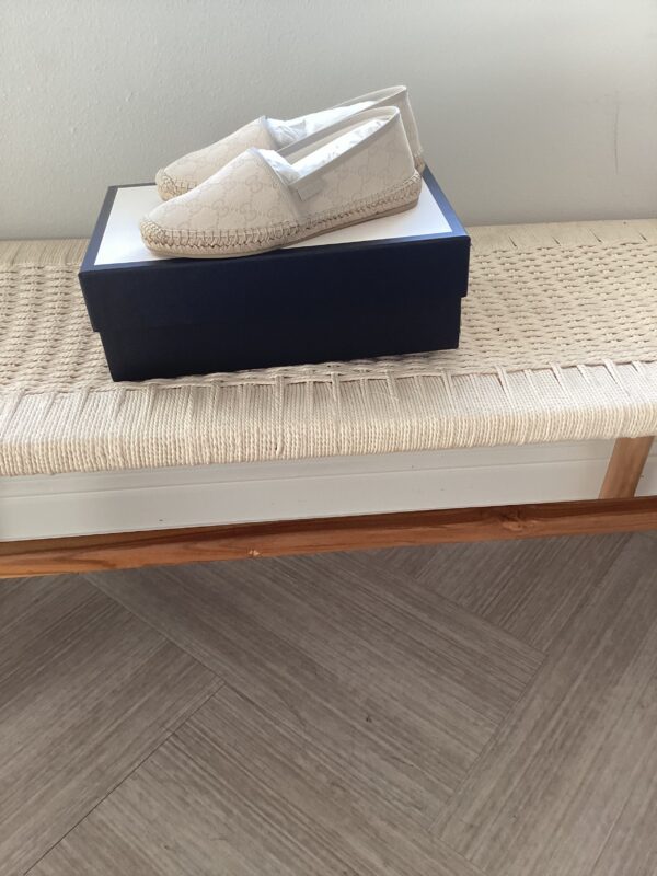 A pair of beige Gucci espadrilles on top of their shoebox, placed on a woven rug beside a wooden bed frame.