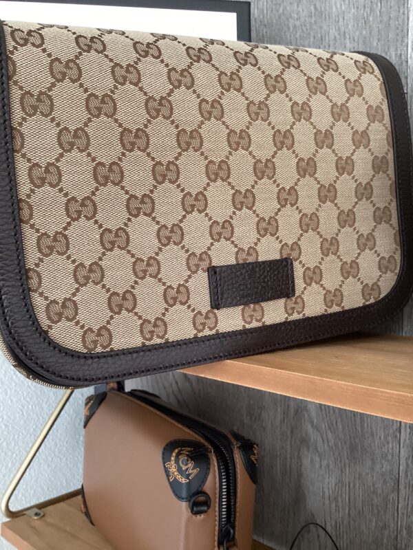 A Gucci Messenger Bag with a brown monogram pattern and black trim, placed on a wooden shelf.