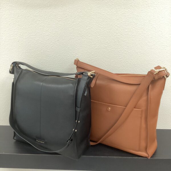 Two Cole Haan bags, one black and one tan, placed side by side on a white surface against a grey wall.
