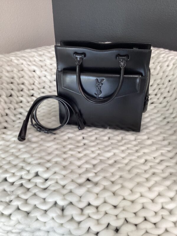 A black YSL Uptown Bag on a white knitted surface, with the strap detached and lying next to it.