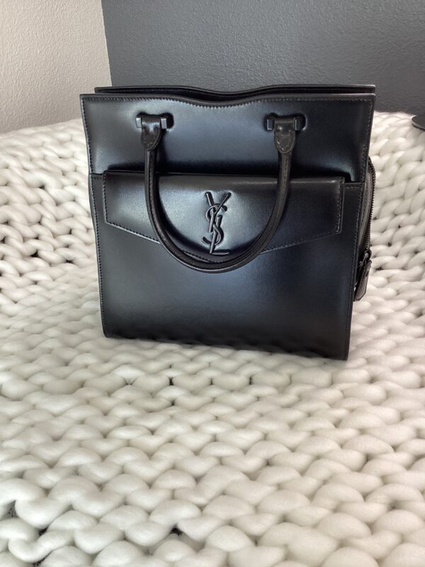 A black YSL Uptown Bag with twin handles, resting on a white textured surface.