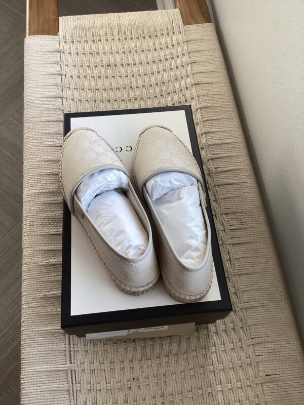 A pair of new Gucci Espadrilles placed on top of their shoebox, which is situated on a woven beige bench.