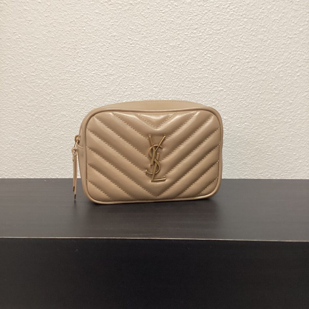 Beige quilted leather YSL 2 Way Belt Bag with a prominent ysl logo on the front, displayed on a dark shelf against a white wall.