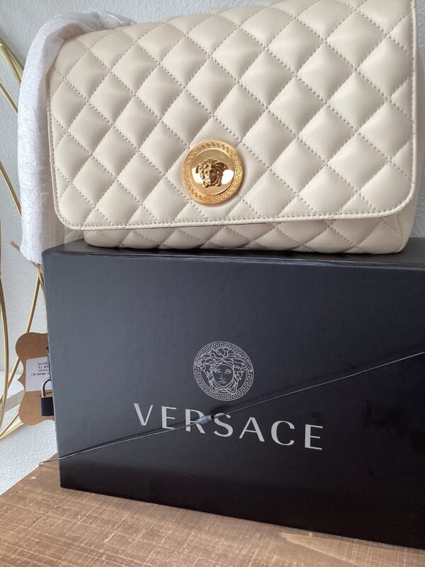 A white quilted purse with a gold emblem rests on a black versace box placed on a wooden surface.