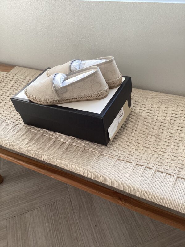 A pair of Gucci Espadrilles with a textured pattern, displayed on an open black shoebox on a woven bench.