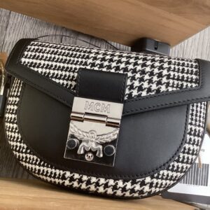 A black and white houndstooth MCM 2-Way Belt bag with a prominent logo clasp, resting on a wooden surface.