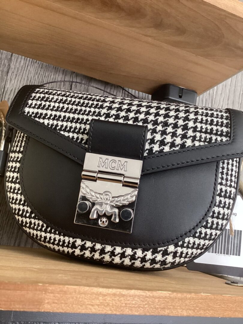 A black and white houndstooth MCM 2-Way Belt bag with a prominent logo clasp, resting on a wooden surface.