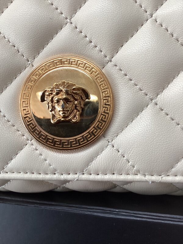 Close-up of a quilted beige handbag featuring a gold medallion with a lion head design surrounded by a greek key pattern.