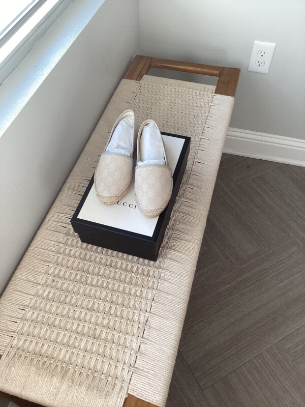 A pair of Gucci espadrilles placed on top of their box on a woven bench near a window in a room with light grey flooring.