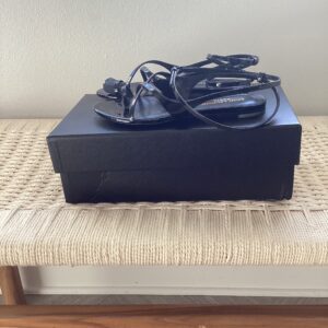 A black Saint Laurent Sandal with cables on top, placed on a beige woven bench against a light grey wall.