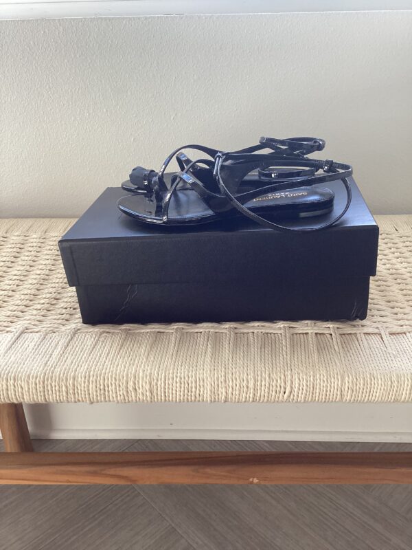 A black Saint Laurent Sandal with cables on top, placed on a beige woven bench against a light grey wall.