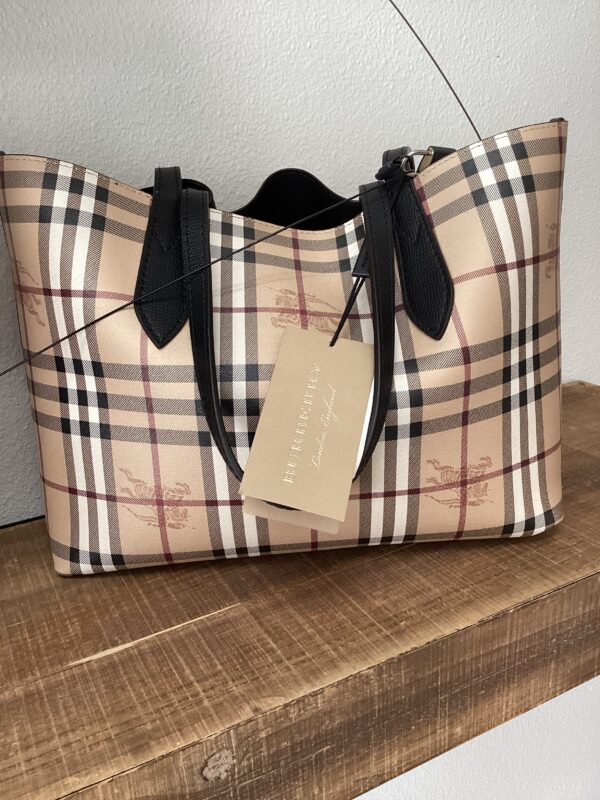 A Burberry (Reversible) Tote with a beige, black, and red plaid pattern, featuring black handles and a brand tag on a wooden surface.