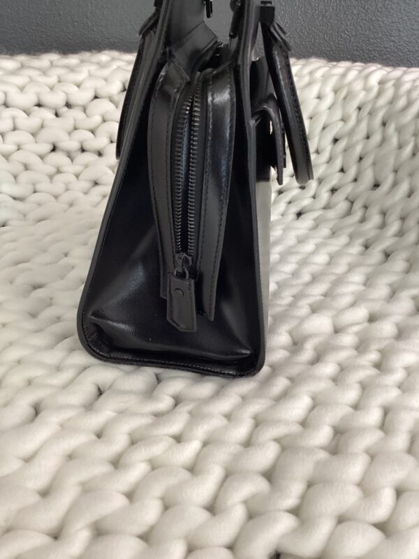 A YSL Uptown Bag in black leather with zipper details, standing on a textured white woven surface.
