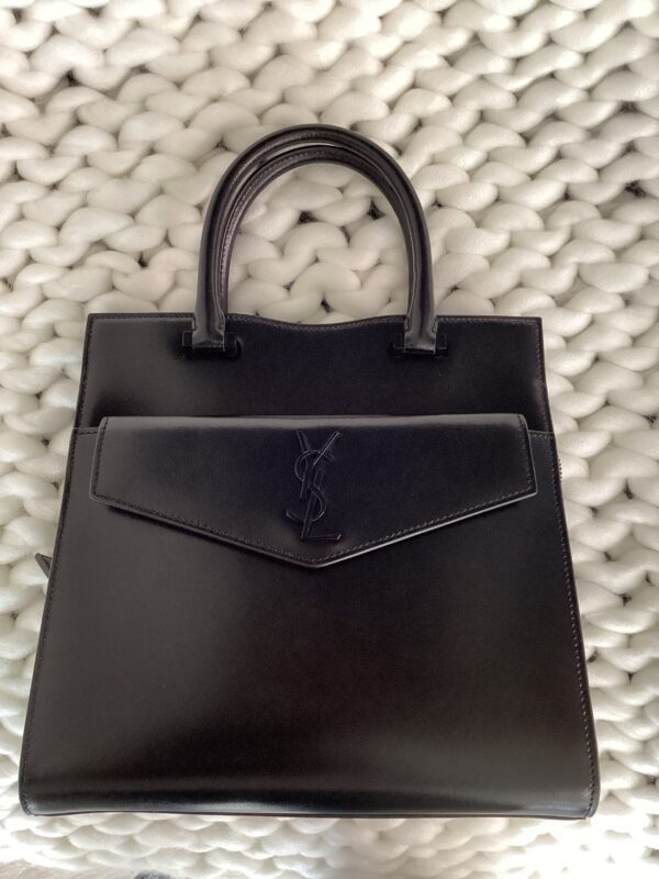 A black YSL Uptown Bag with a top handle, resting on a white woven background.