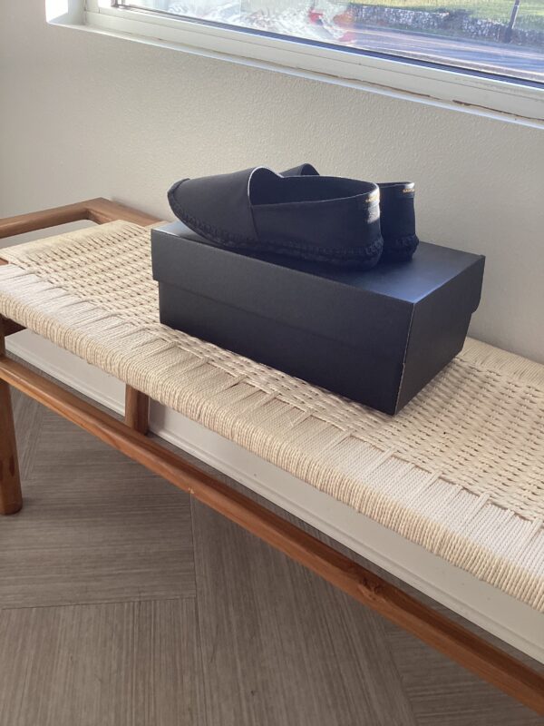 A pair of YSL Espadrilles placed neatly on top of a shoebox, which is sitting on a woven beige bench by a window.