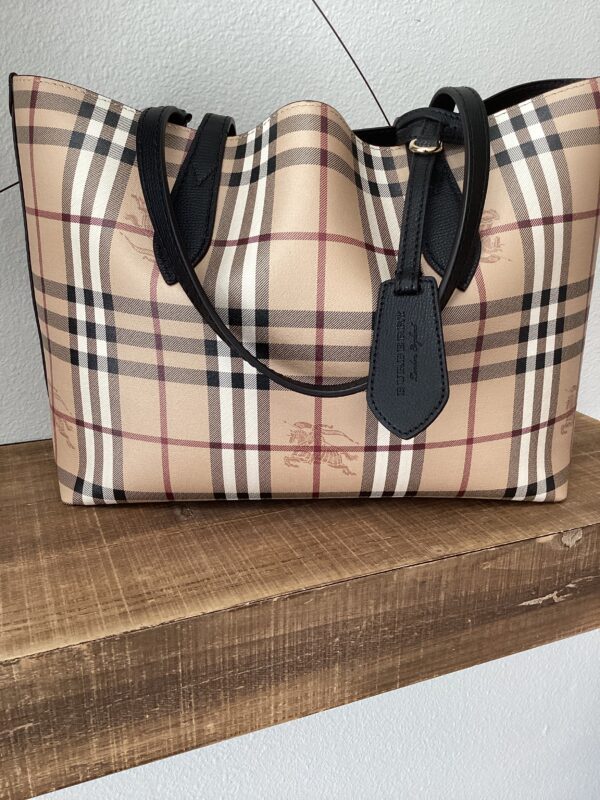 A beige Burberry (Reversible) tote bag with a checkered pattern and black handles, displayed on a wooden shelf.