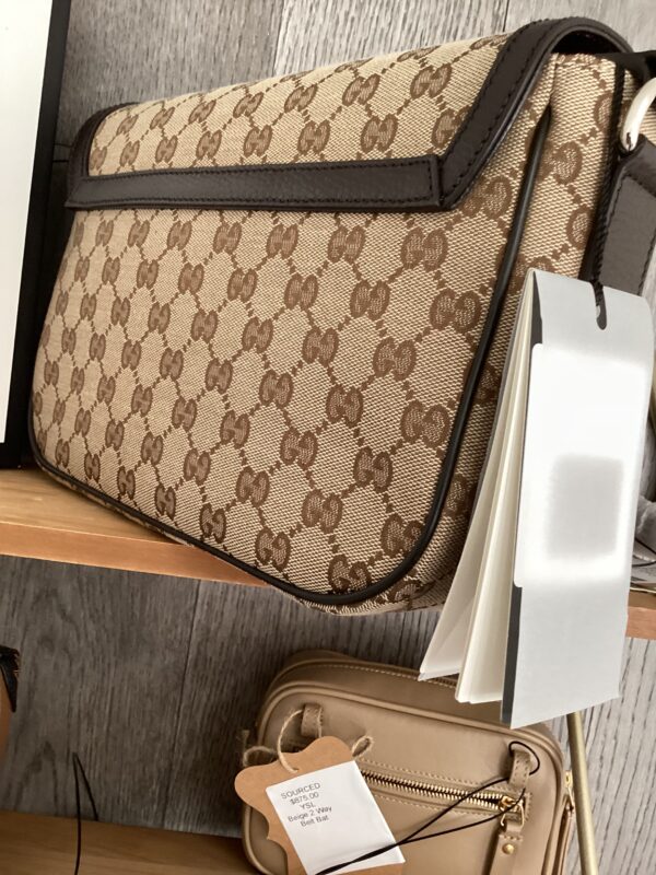 A Gucci Messenger Bag with a monogram pattern, displayed on a wooden shelf beside a white electronic device and a hanging tag.