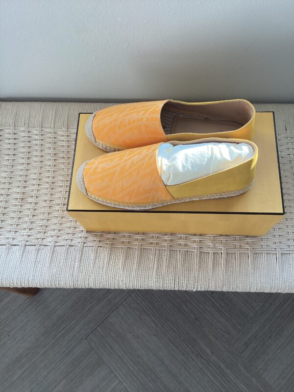 A pair of Fendi Yellow Espadrilles with orange textured uppers and metallic gold toes, displayed on a gold box atop a woven bench.