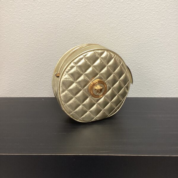 A small, round, VERSACE Gold Bag with a zipper and a logo emblem, placed on a grey surface against a light blue wall.