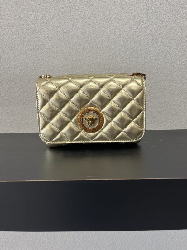 VERSACE Gold quilted shoulder bag with a metallic logo displayed on a shelf against a grey wall.