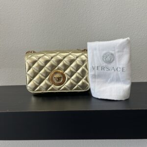 VERSACE Gold quilted handbag with a circular logo next to a folded white cloth embroidered with the VERSACE logo, positioned on a black shelf.
