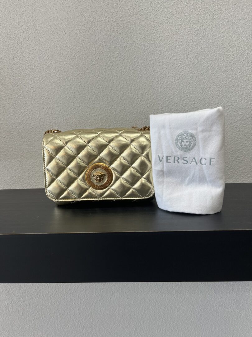 VERSACE Gold quilted handbag with a circular logo next to a folded white cloth embroidered with the VERSACE logo, positioned on a black shelf.