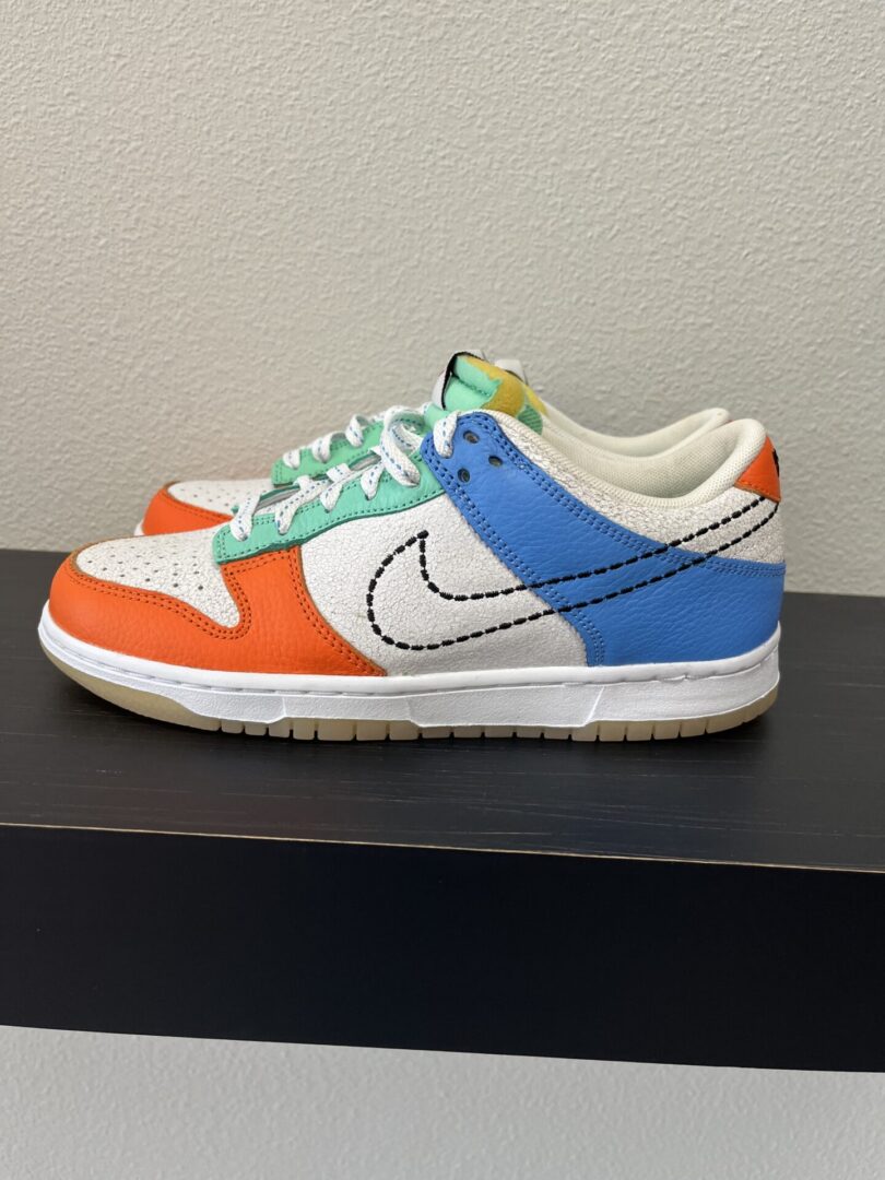 A colorful sneaker with orange, blue, and white panels and green laces on a simple black shelf.