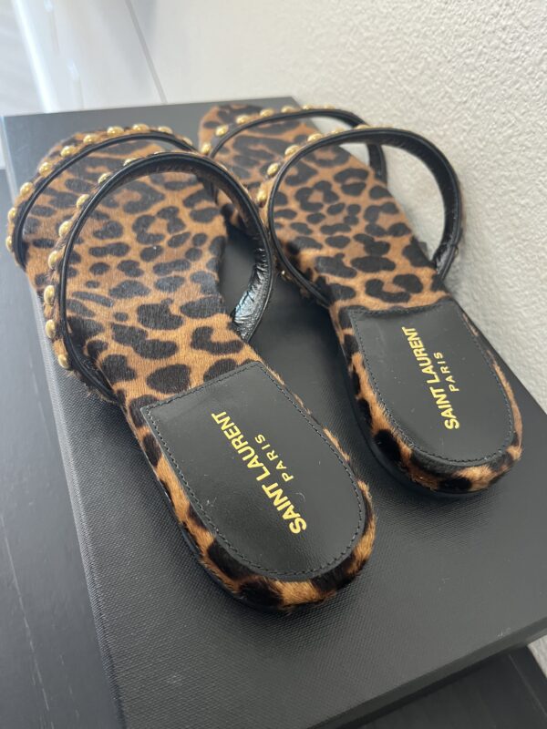 A pair of YSL Kiki Stud Sandals with leopard print and studded straps, displayed on a black surface.