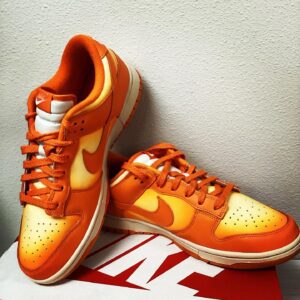 A pair of orange Magma Dunk Low sneakers placed on a white Magma Dunk Low shoebox against a textured grey background.