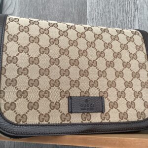 A beige Gucci Messenger Bag with a repeating logo pattern, featuring a black leather label on the front, resting on a wooden surface.