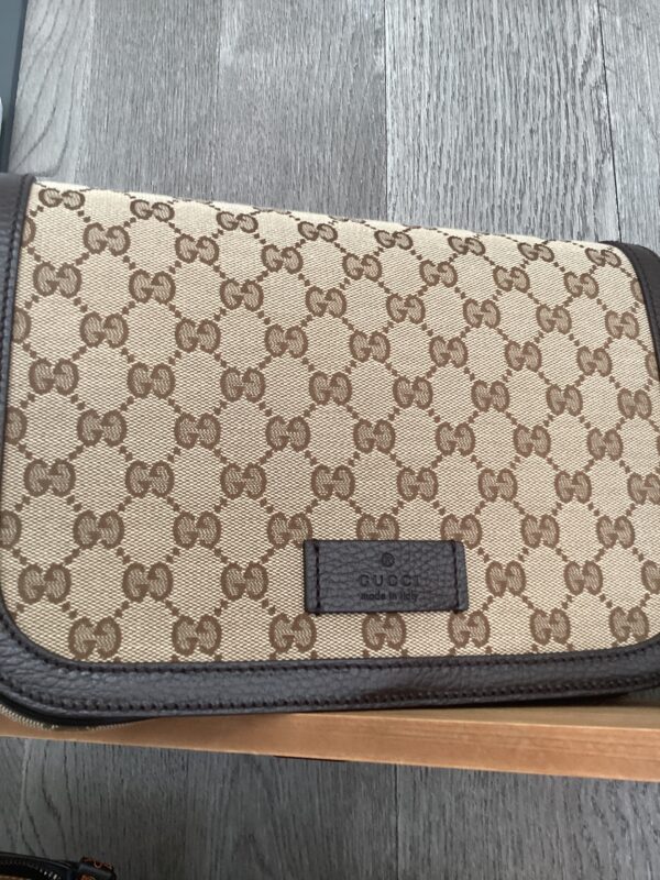A beige Gucci Messenger Bag with a repeating logo pattern, featuring a black leather label on the front, resting on a wooden surface.