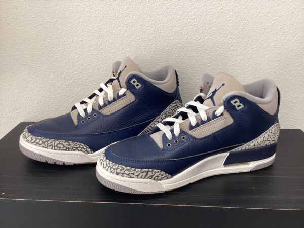 A pair of blue and grey air jordan 3 sneakers displayed on a black shelf against a white wall.