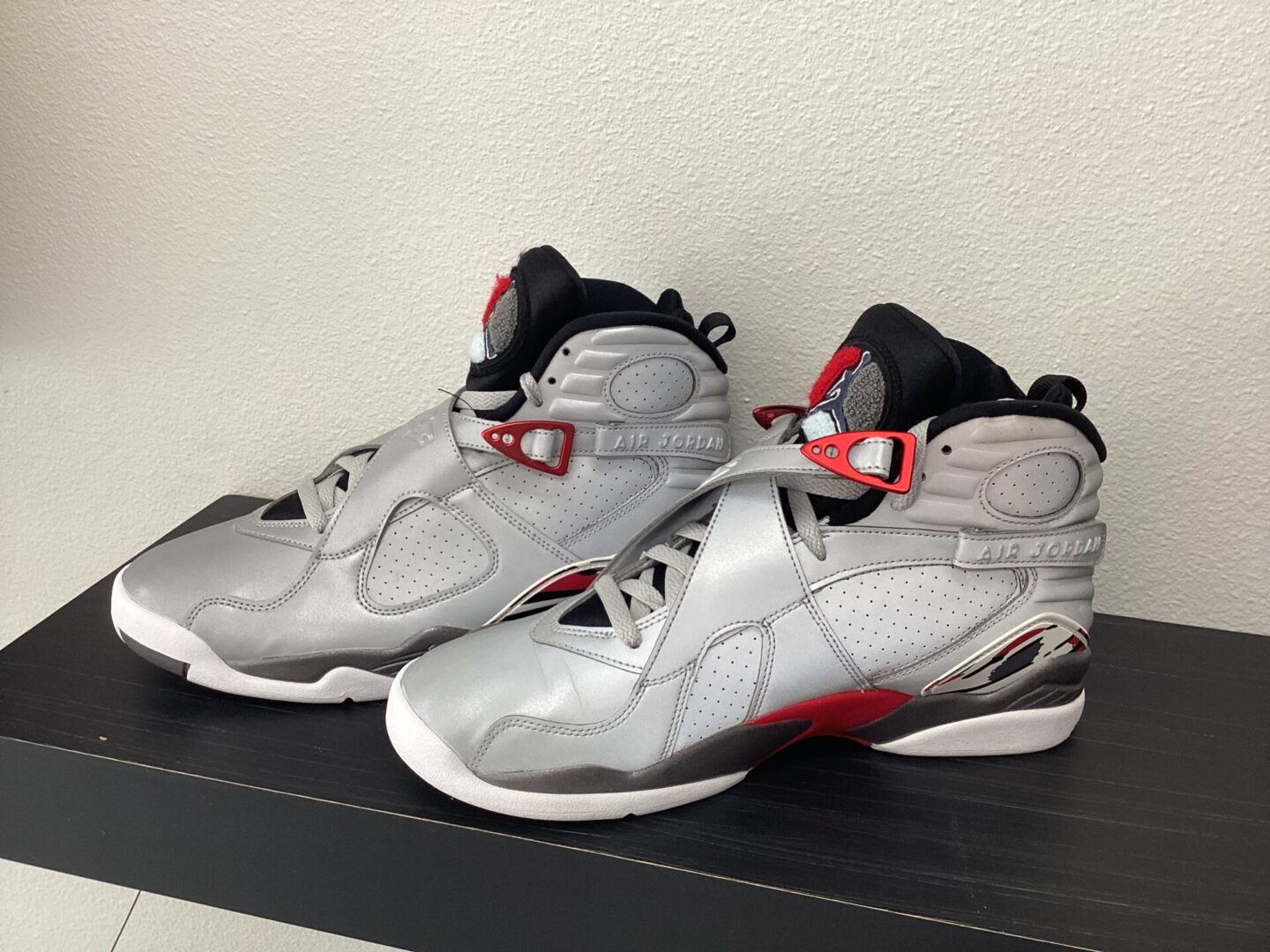 A pair of grey and red high-top sneakers with "Pre owned-Jordan 8" text displayed, resting on a black shelf against a white wall.