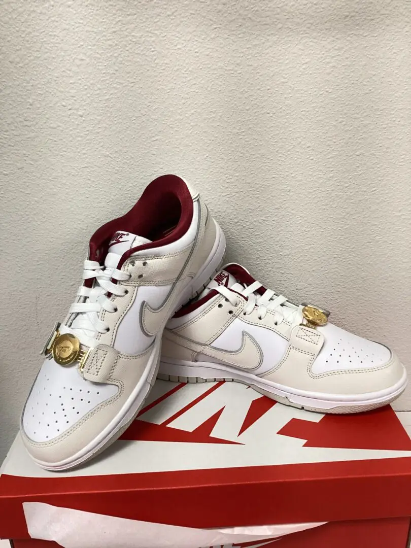A pair of white W Nike Dunk Low sneakers with gold accents and a small gold watch attached to one shoe, displayed on a red nike shoebox.