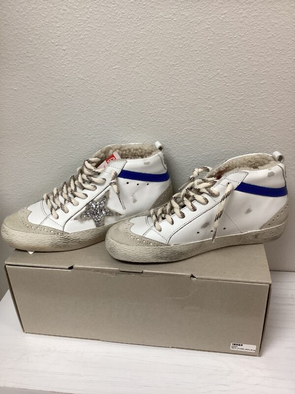 A pair of worn white high-top sneakers with glitter details and blue accents, displayed on top of a cardboard shoebox against a plain wall.