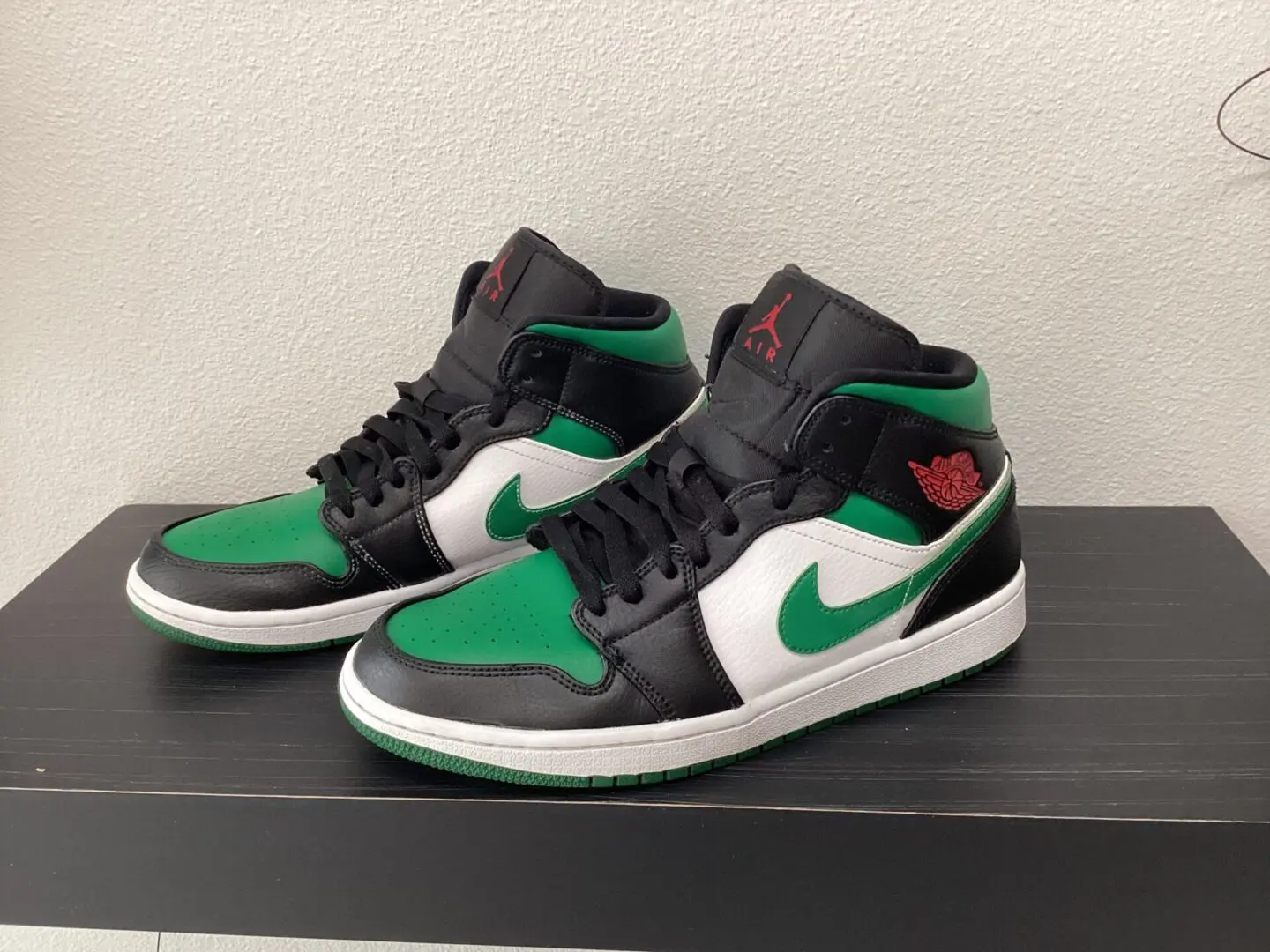 A pair of black and green Pre-owned Jordan 1 Mids sneakers with white nike swoosh logos displayed on a black shelf.