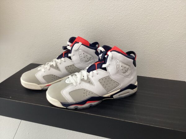 A pair of Pre-Owned Jordan 6 with white, red, and navy blue accents displayed on a black shelf against a grey wall.