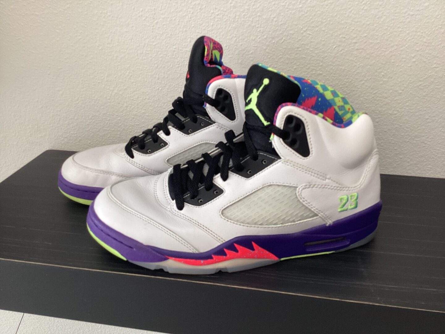 A pair of Pre owned- Jordan 5 Prince sneakers with a white and gray upper, colorful inner lining, and a purple and red sole on a black shelf.
