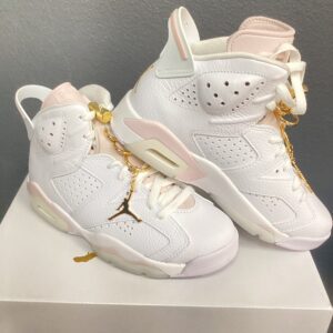 A pair of white and pink **Jordan 6 Retro white/metallic gold** sneakers with gold accents displayed on a shelf.